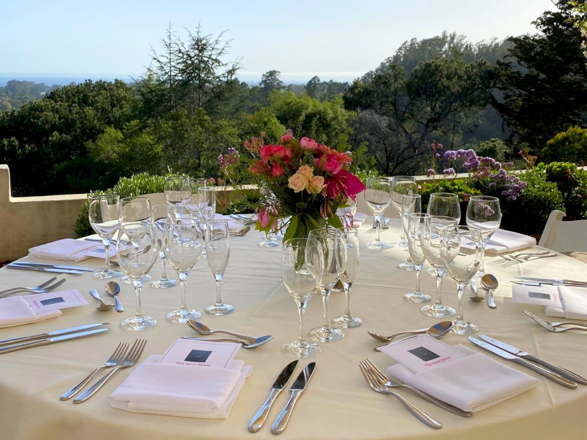 A beautifully set round table with glasses, cutlery, and napkins overlooks a scenic landscape, adorned with a centerpiece of vibrant flowers.