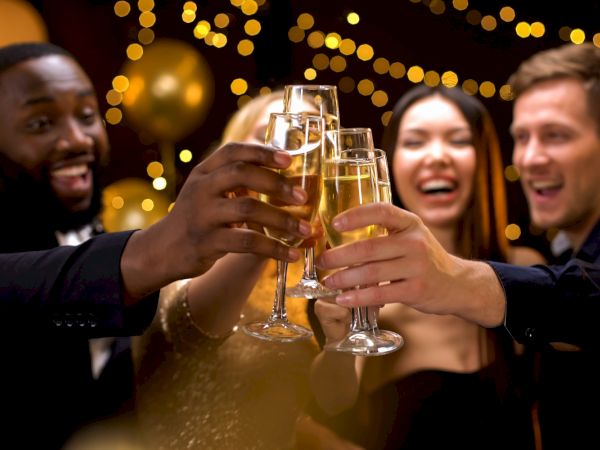 A group of smiling people dressed up, clinking champagne glasses in celebration, with festive lights and balloons in the background.