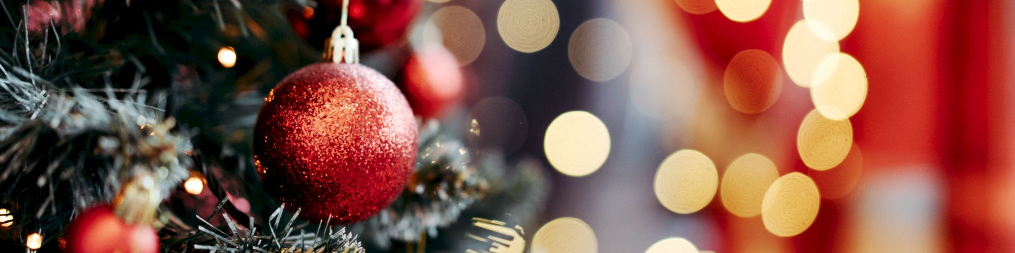 A decorated Christmas tree with red baubles and festive lights. The background shows out-of-focus, warm bokeh lights.