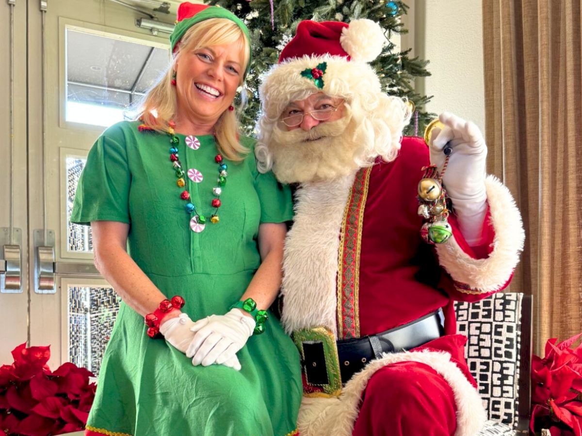 A person dressed as Santa Claus sits next to someone in an elf costume in front of a Christmas tree, surrounded by festive decorations.