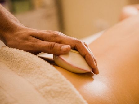 The image shows a person receiving a hot stone massage on their back, with a towel and the masseuse's hand holding a large stone.