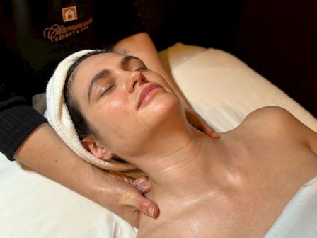 A person is receiving a neck massage while lying on a massage table, eyes closed, with a towel wrapped around their head.