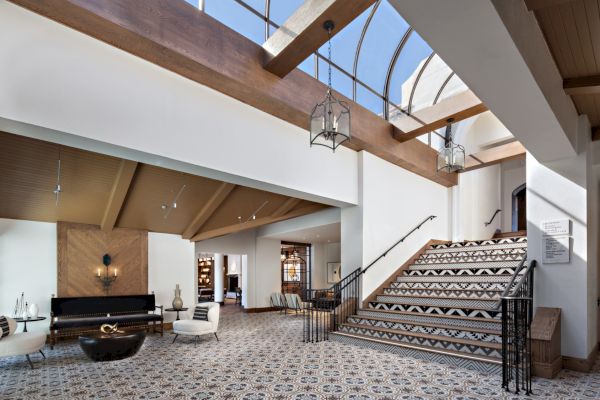 A spacious, modern lobby with patterned flooring, a staircase, natural light, furniture, and decorative lamps under a skylight ceiling.