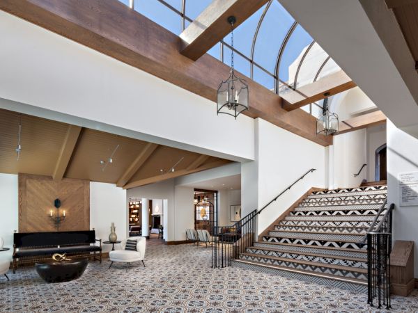 A spacious, modern lobby with patterned flooring, a staircase, natural light, furniture, and decorative lamps under a skylight ceiling.