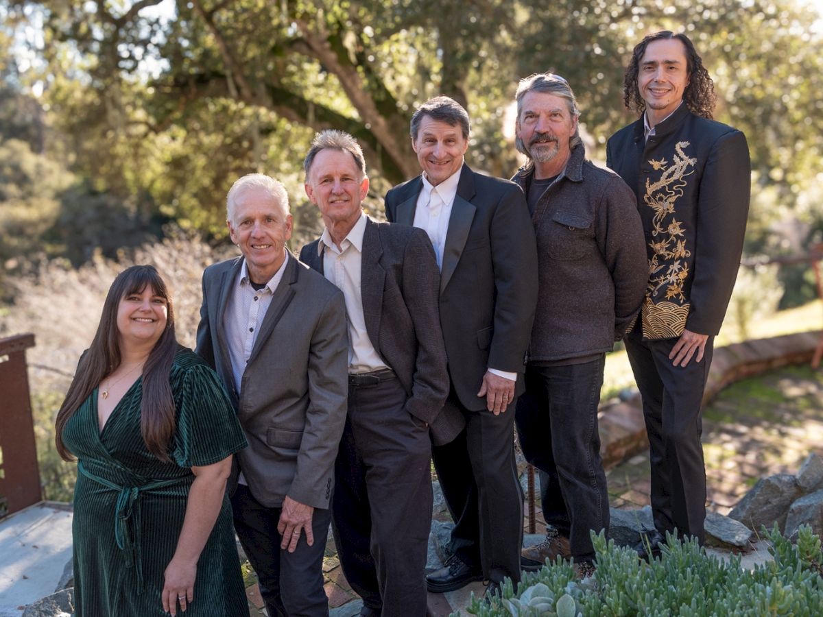 A group of six people dressed in formal and semi-formal attire stand outdoors, surrounded by greenery, posing for a photo.