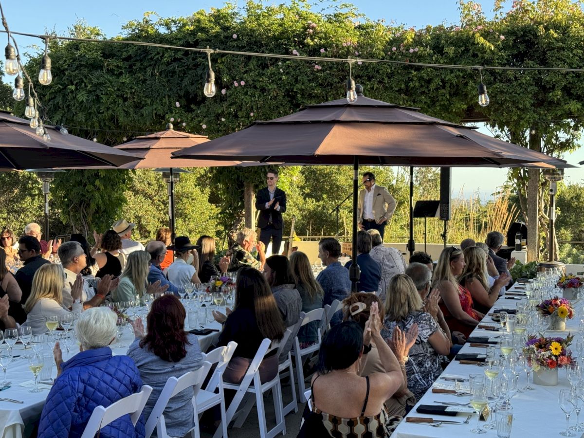 Outdoor gathering with people seated at long tables, listening to a speaker under umbrellas. Trees and string lights in the background.