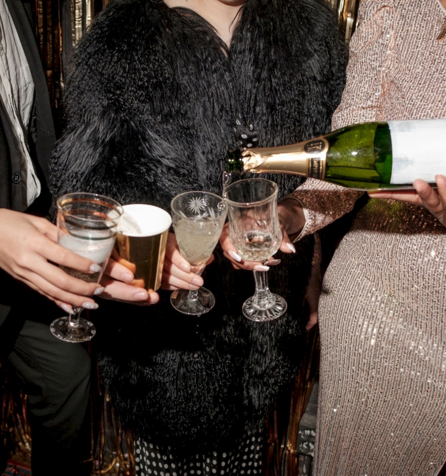 People at a party pouring champagne into glasses, dressed in fancy attire, with a festive atmosphere.