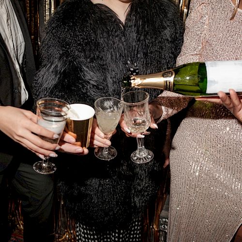 People at a party pouring champagne into glasses, dressed in formal attire with festive decorations in the background.