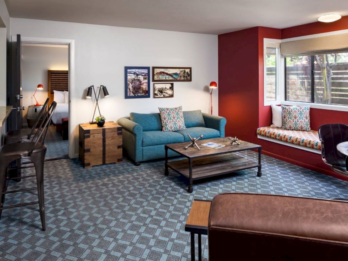 A cozy living room with a blue sofa, patterned carpet, red accent wall, and large window. A bedroom is visible through an open door.