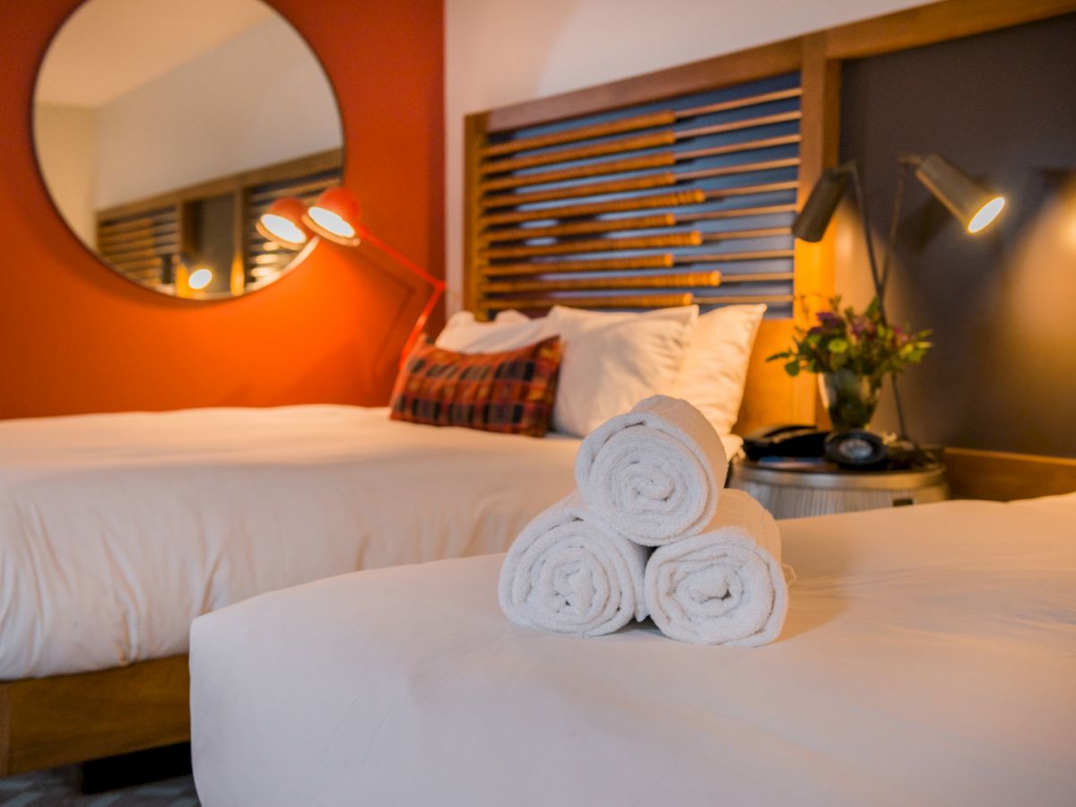 The image shows a neatly arranged hotel room with two beds, rolled towels, stylish lamps, a wooden headboard, and a round mirror above the beds.