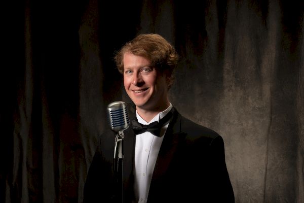 A man in a tuxedo stands in front of a vintage microphone with a dark curtain backdrop, smiling at the camera.