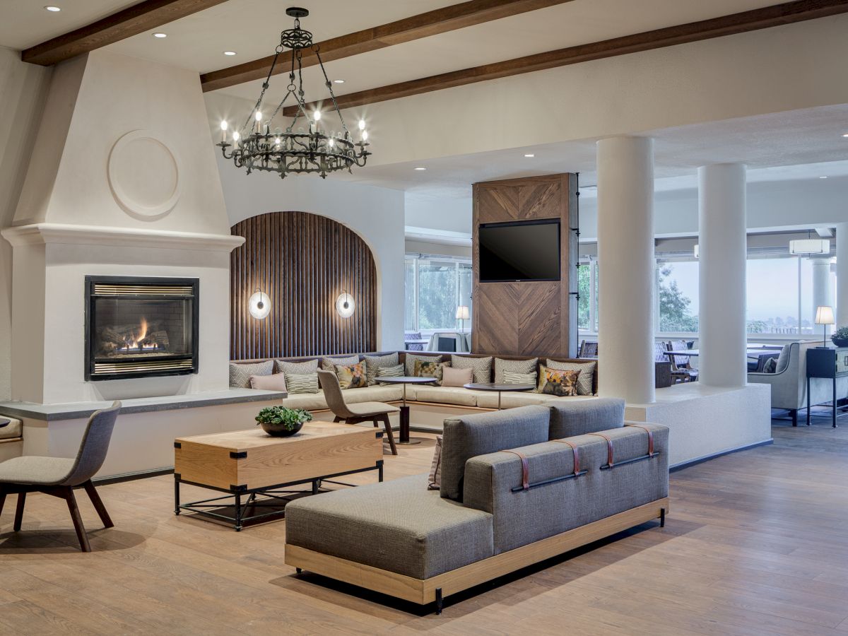 A modern lounge area with cozy seating, a fireplace, chandelier, and a mounted TV, featuring large windows for natural light.