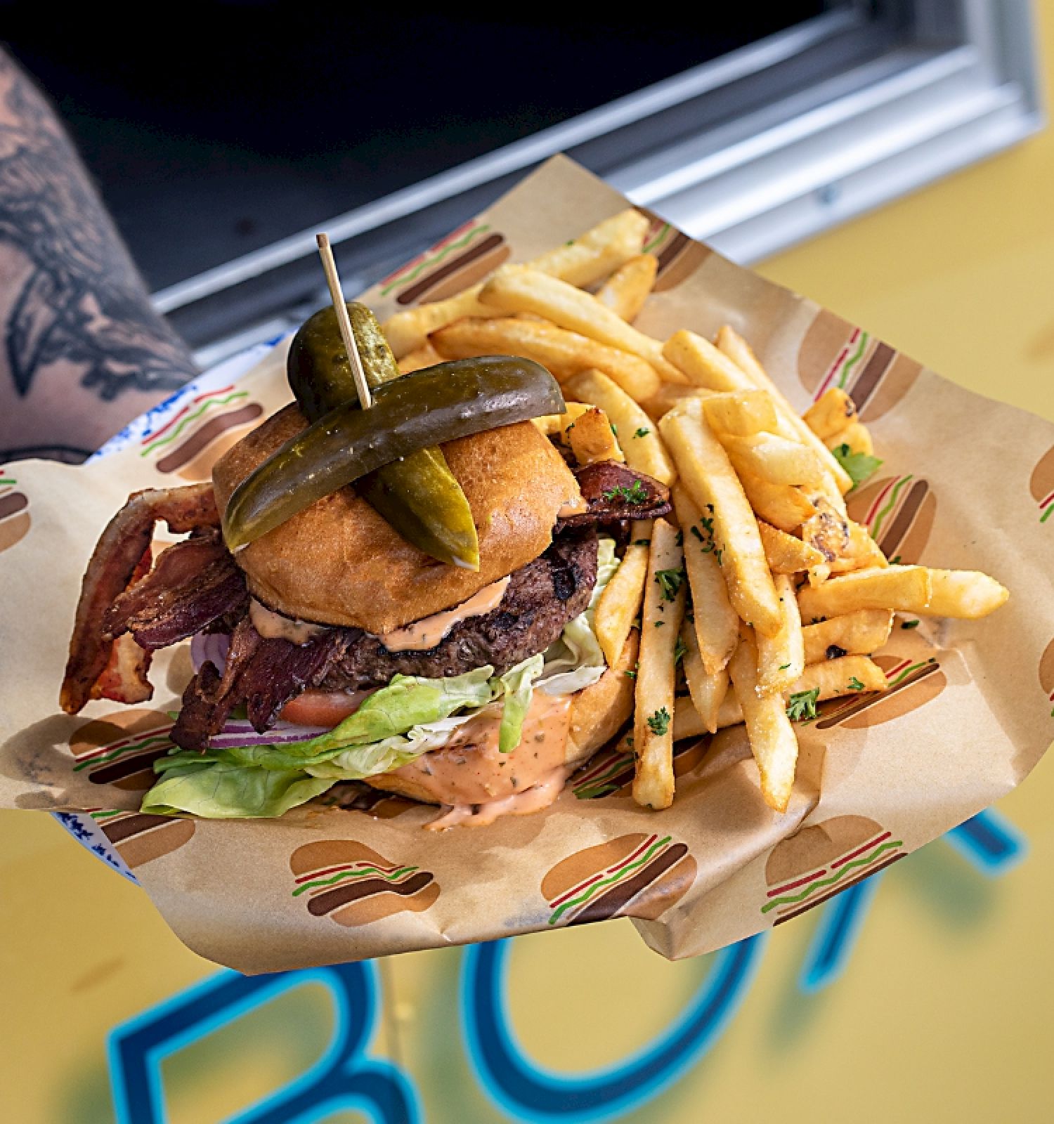 A tattooed arm holds a burger with pickles and fries on a paper tray in front of a yellow food truck.