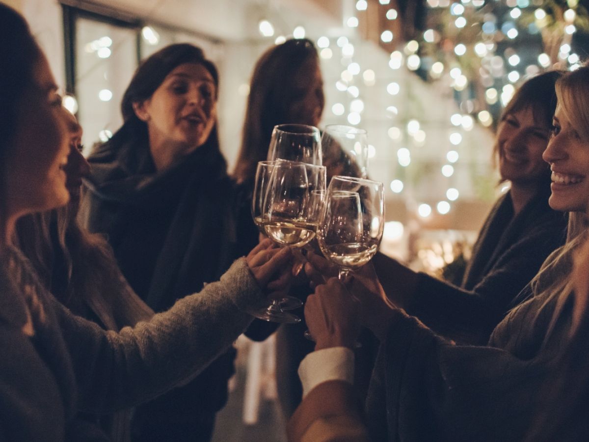 A group of people is cheerfully clinking wine glasses together in a warmly-lit, festive setting with string lights in the background.