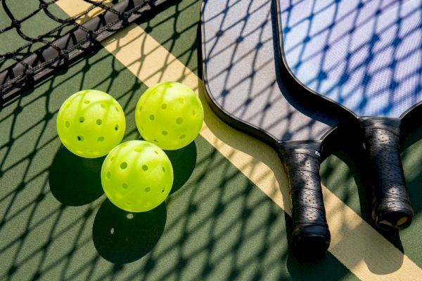 The image shows pickleball paddles, bright yellow pickleballs, and a net on a court.