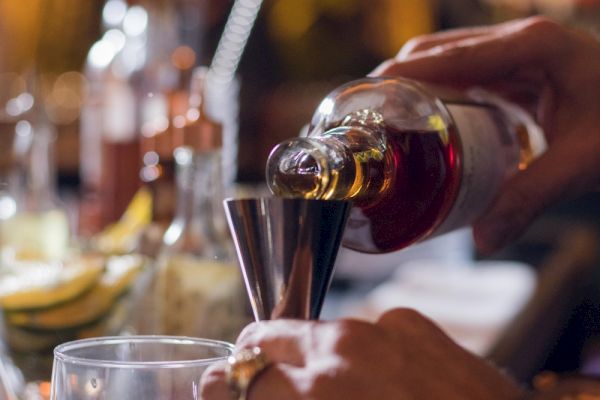 A person pours a brown liquid from a bottle into a jigger, surrounded by bar tools and sliced lemons on a counter.