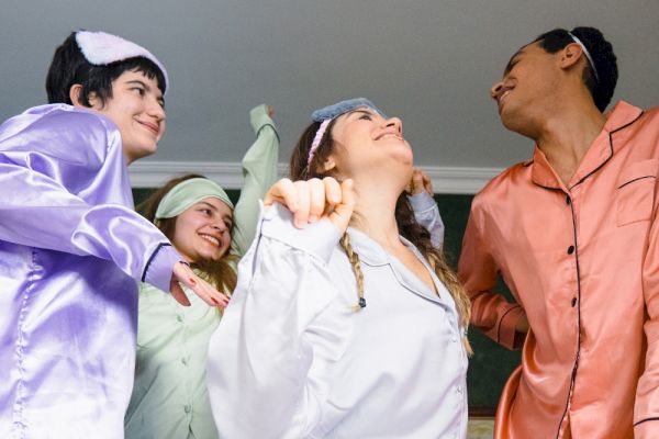 Four people in colorful pajamas are joyfully dancing together, each wearing a sleep mask on their heads, in a fun indoor setting.