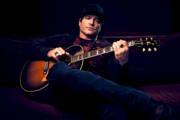 A person is sitting on a couch, playing an acoustic guitar, wearing a dark hat and clothing against a dark background.