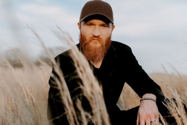 A person with a beard and cap sits in a field of tall, dry grass under a blue sky, wearing a black shirt, looking towards the camera.