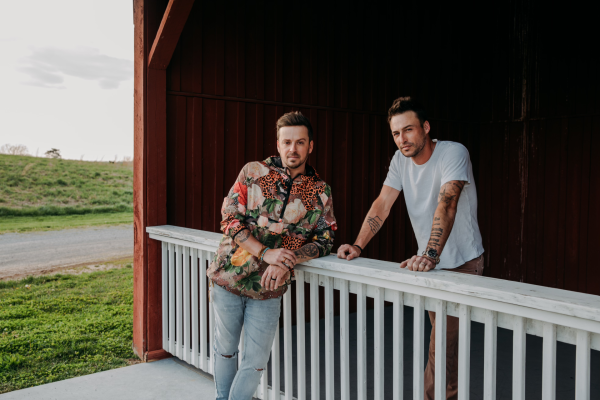 Two people are leaning on a white railing in front of a red building, with grass and a road in the background.