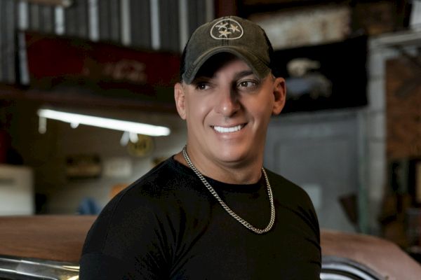A person in a cap and black shirt with a chain necklace smiles in an industrial setting with a car in the background.