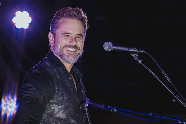A man with a beard stands on stage smiling, near a microphone, under bright lighting.