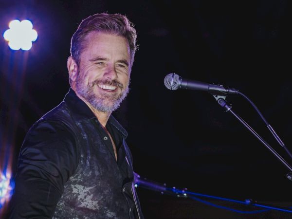 A man with a beard stands on stage smiling, near a microphone, under bright lighting.