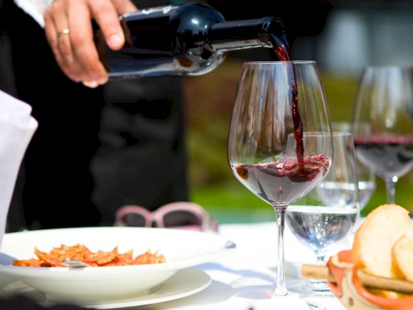 A hand pours red wine into a glass at a table with plates of food and bread, with another glass and sunny outdoor light nearby.