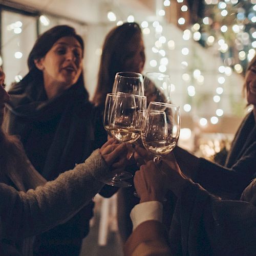 A group of people smiling and raising glasses in a toast, surrounded by festive lights in a cozy, dimly lit setting.