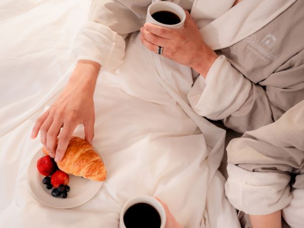 Two people in robes enjoy breakfast in bed, featuring coffee and a plate with a croissant and berries.