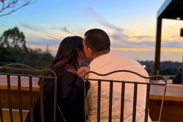 A couple is sitting closely on outdoor chairs, sharing a moment during a sunset with a scenic view in the background.