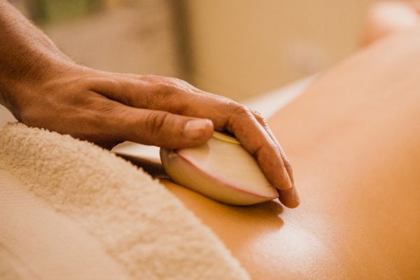 The image shows a person receiving a hot stone massage on their back, with a towel and the masseuse's hand holding a large stone.