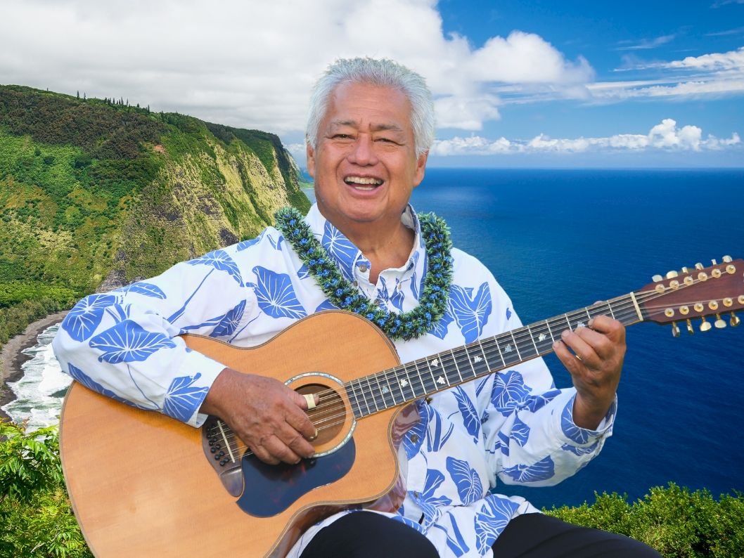 A smiling man playing an acoustic guitar on a cliffside with blue ocean, green hills, and a bright sky behind him, wearing a blue Hawaiian shirt.