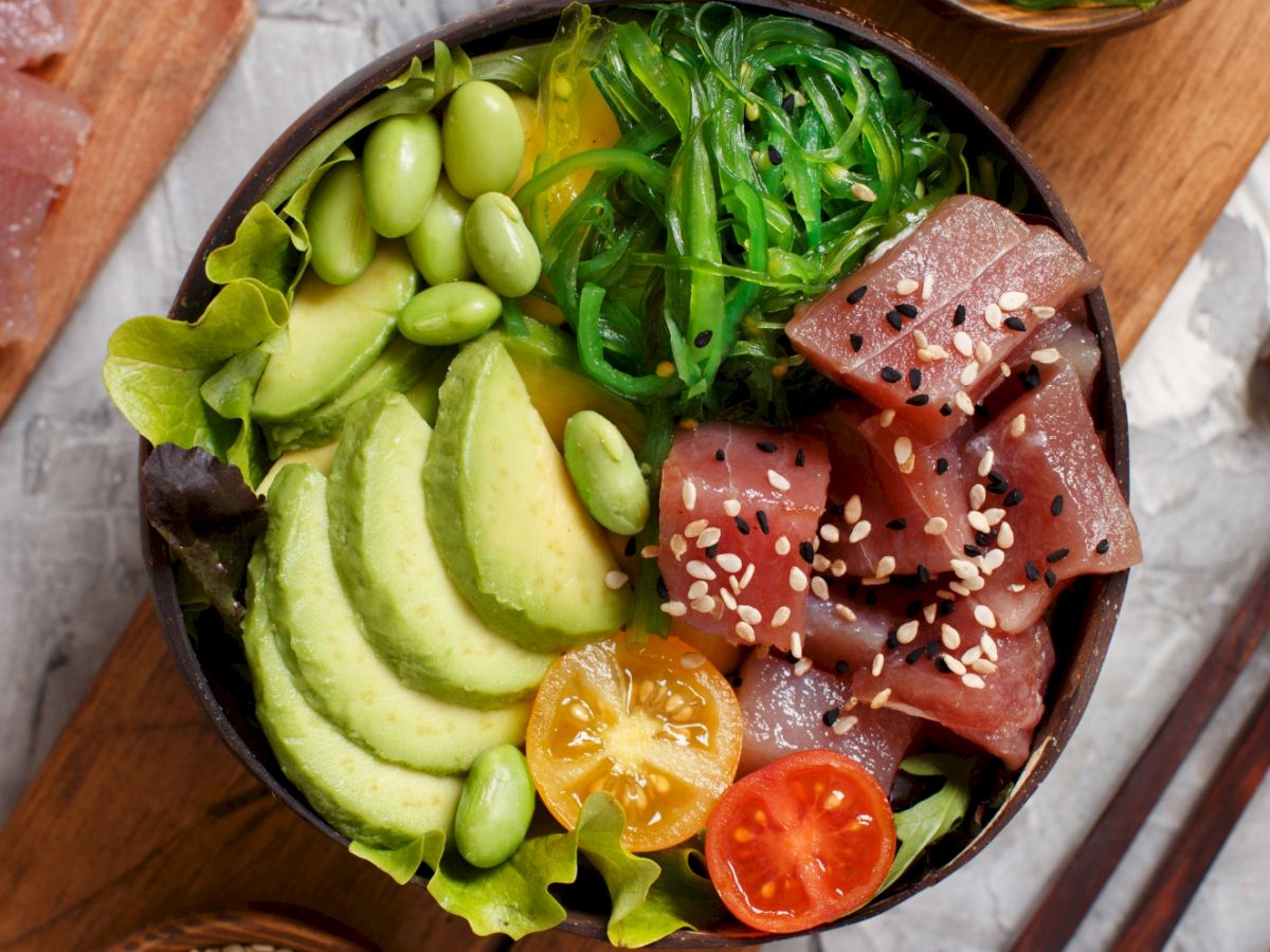 A poké bowl with tuna, avocado, edamame, seaweed salad, tomatoes, lettuce, and sesame seeds, arranged neatly in a bowl.