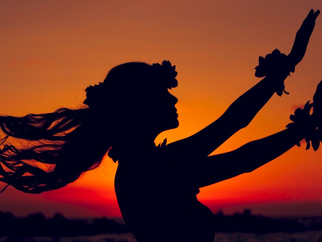 A silhouetted person with arms raised against a vibrant sunset, wearing a flower headband and wrist garlands, dancing by the water.