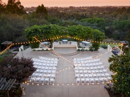 An outdoor event setup with rows of white chairs, string lights, surrounded by greenery and a scenic background at sunset.