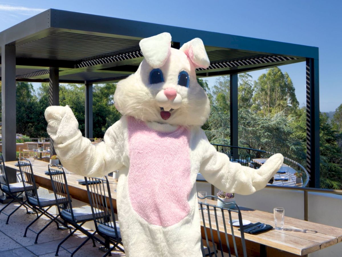 A person in an Easter Bunny costume is posing on a sunny restaurant patio with tables set for dining.