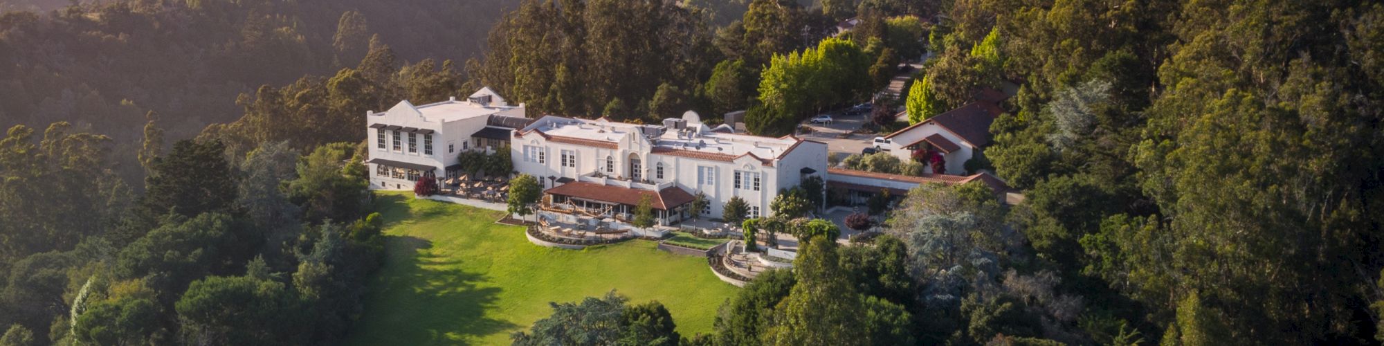 An aerial view of a large, luxurious mansion surrounded by lush greenery and hills with a well-maintained garden and trees.