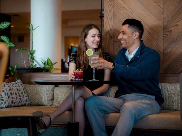 A couple sits on a cozy couch at a bar, sipping drinks and smiling at each other, enjoying a night out together.
