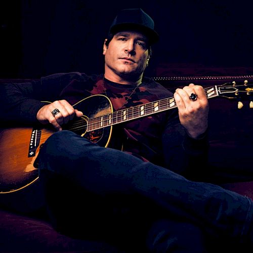 A person lounging on a sofa, holding an acoustic guitar, wearing casual clothing, and a cap, set against a dark background.