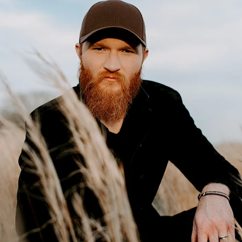 A person with a beard and cap is sitting in a field of tall grasses, looking directly at the camera under a partly cloudy sky.