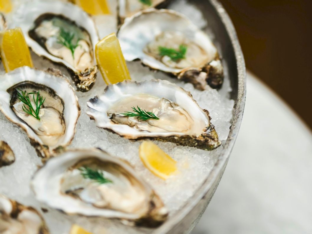 Oysters on ice garnished with dill and small lemon pieces, presented in a round platter.
