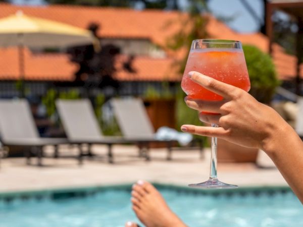 A hand holds a colorful drink by a pool, with lounge chairs and umbrellas in the background.