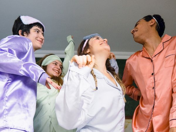 Four people in colorful pajamas are joyfully dancing together, each wearing a sleep mask on their heads, in a fun indoor setting.