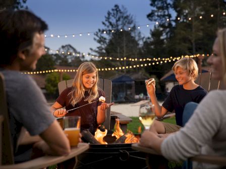 A group of four people sits around a fire pit, enjoying roasted marshmallows and drinks, with string lights overhead and trees in the background.