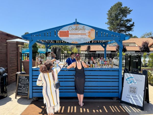 A blue outdoor cocktail stand with two customers ordering drinks in a sunny setting. There's signage and various bottles on display.