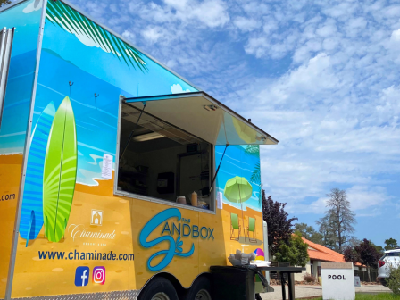 A colorful food truck named "Sandbox," with surfboards painted, set near a pool area. The sky is partly cloudy.
