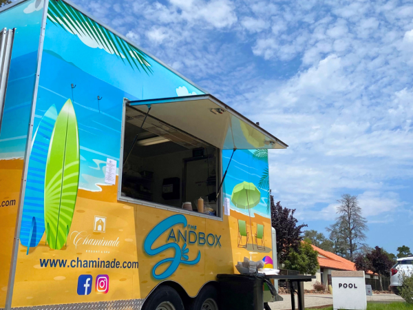 A colorful food truck named "Sandbox," with surfboards painted, set near a pool area. The sky is partly cloudy.