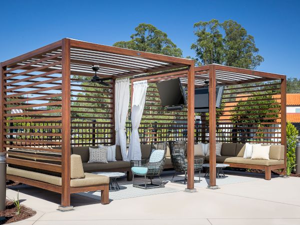 A modern outdoor patio area with wooden pergolas, seating, tables, and a TV. Surrounded by trees and landscaped greenery, under a clear sky.