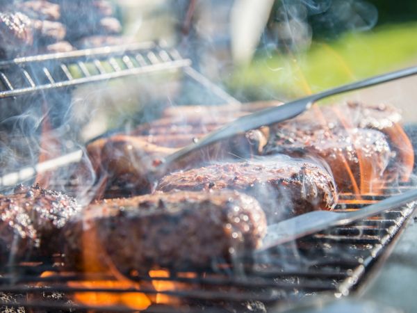 Grilled burgers and sausages on a barbecue with tongs flipping meat, surrounded by smoke and flame, perfect for a summer cookout.